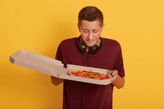 Close up portrait of handsome guy wearing burgundy t shirt smelling fresh pizza from carton box and gets pleasure, standing isolated over yellow background, young man being ready to eat junk food.