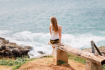 A girl sits on a bench and looks at the beautiful sea. Dreamy blonde and traveler in a white tank top and black shorts. Tropical Island Sri Lanka. - Powered by Adobe