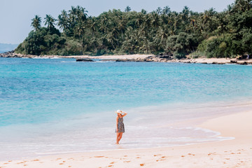 Fototapeta na wymiar A girl walks along the sea. The blonde in a colored dress and a straw hat rejoices in the waves. Pink sand beach and beautiful palm trees in Sri Lanka.