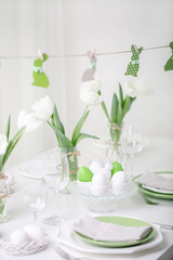 Decor and table setting of the Easter table with white tulips and dishes of green and white color. Easter decor in the form of Easter bunnies  green color with white polka dots.