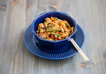 Teriyaki chicken with noodles and vegetables stir fry in a bowl, wooden background. Udon noodles, teriyaki chicken, vegetables, chopsticks. Closeup. Asian style dinner.