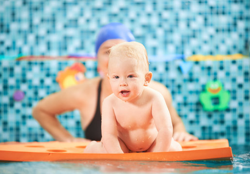 Horizontal picture of a cute little child spending fun time in swimming pool. A boy is crawling on a floating mat, mother is behind him supporting. Concept of active leisure