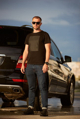 Fototapeta na wymiar Portrait of a successful man wearing sunglasses, black T-shirt, dark blue jeans and black sneakers is standing next to his black car, full length