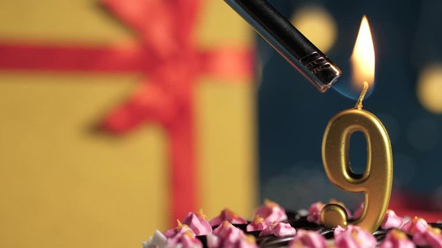 Birthday cake number 9 golden candles burning by lighter, blue background gift yellow box tied up with red ribbon. Close-up and slow motion