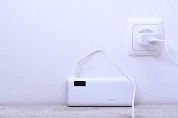 White phone power bank plugged in the socket on the wall for charging on wooden background