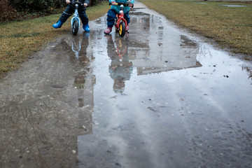 Zwei Kinder fahren mit dem Laufrad bei Regen durch eine Pfütze