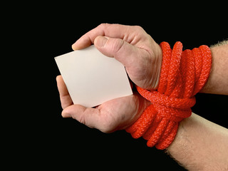 Hands tied with a rope, on a black background. Tied hands are holding a note with the inscription....