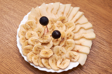 Sliced banana, apple, plum on a white round plate on a wooden table. Fruit on a plate. The image on white background. View from the top.