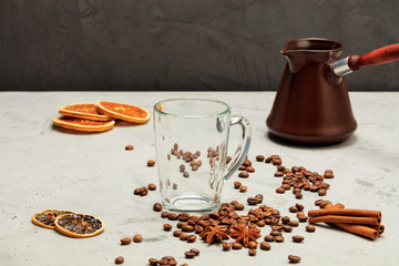 A transparent glass cup stands on a background of gray concrete with coffee beans, dried orange and lemon, anise, cinnamon and coffee cezve in blur.
