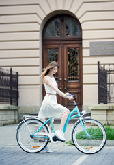 Naklejka na ściany i meble Lovely girl in a light dress and sneakers on a blue retro bike against the background of an old building with a beautiful door. Bicycle rides in summer afternoon outdoors
