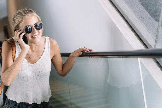 Girl with sunglasses and a bag hanging from her shoulder talking on her mobile phone while she smiles in an escalator