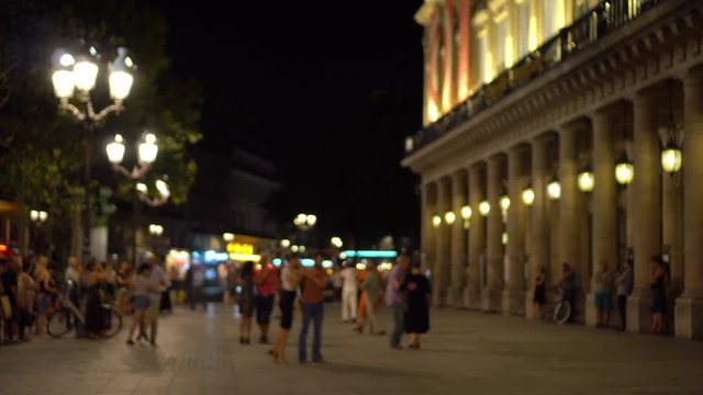 Parisians and tourists dance elegantly in one of the squares of Paris. Facade of the building is decorated with columns, tree branches, lights of passing cars in the background. Outdoor, summer night.