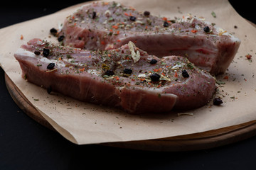 Pink raw pork steaks lie on a wooden board in spices and green dill. Appetizing meat. On kraft paper and a black background.
