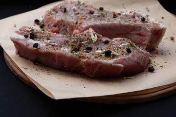Pink raw pork steaks lie on a wooden board in spices and green dill. Appetizing meat. On kraft paper and a black background.