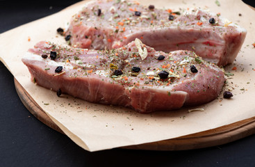 Pink raw pork steaks lie on a wooden board in spices and green dill. Appetizing meat. On kraft paper and a black background.