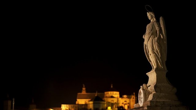 Pan shot left to right of Saint Raphael statue at night on the Roman Bridge of Cordoba. View of Mosque-cathedral, Catedral de Cordoba behind an angel statue on the roman bridge