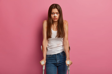 Offended woman with bruises and broken nose, being seriously injured, faces disaster, misfortune and tragedy, stands on crutches, poses against pink background. Innocent victim with severe traumas