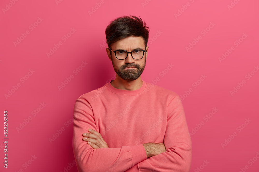 Wall mural Disappointed displeased bearded European man crosses arms and presses lips, expresses resentment and anger, has bad mood, looks with serious displeased expression, isolated over pink background.