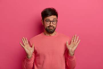 Naklejka premium Portrait of bearded man raises palms, shows beig not involved or guilty, gestures in studio, has confused look after doing something wrong, feels sorry, wears pink jumper. I couldnt handle it