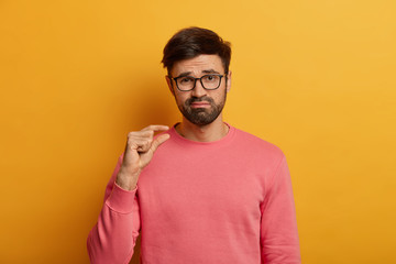 Calm bearded male model shapes small item, frowns face from dislike and regret, demonstrates size...