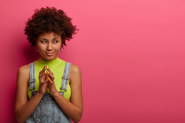 Portrait of pleased dark skinned woman has tricky plan in mind, steeps fingers and looks aside, wears denim overalls, isolated over pink studio wall, empty space, has intention to do something