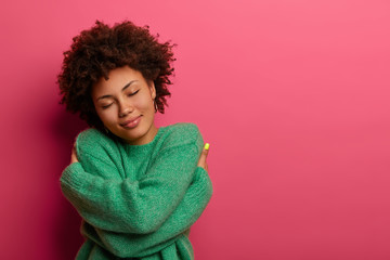 Cheerful young romantic woman expresses self love and care, tilts head and smiles gently, wears green oversized jumper, embraces own body, closes eyes, stands in studio against pink background - Powered by Adobe