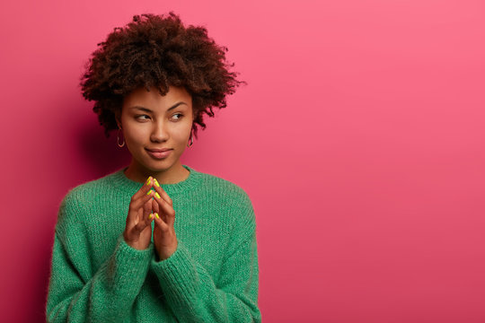 Serious curly haired woman steepls fingers, has cunninng expression, says everything goes by evil plan, intention to do something, wears green sweater, models over rosy wall, empty space area