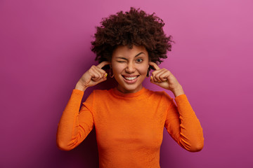Cheerful curly haired woman feels relieved, plugs ear holes with index fingers, hears no sound or noise, shows white teeth, wears casual clothing, poses indoor. Body language, reaction, attitude
