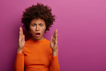 Displeased emotional woman with Afro hairstyle shapes someting big, impressed badly by huge item, raises palms, demonstrates size of object and claims, wears casual orange jumper, isolated on purple