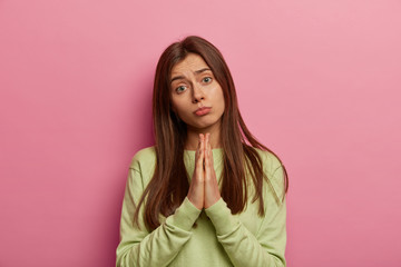 Portrait of imploring girl tilts head, purses lips, looks sadly at camera, makes praying gesture, asks for help and support, begs boyfriend for apologize, wears casual sweater, isolated on pink wall