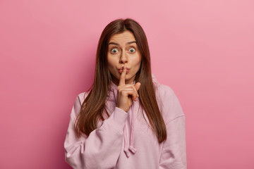 Young wondered woman makes silence sign, tells private information, presses fore finger on lips keeps secret or gossips about something wears long sleeved sweatshirt poses indoor. Hush, shh, be silent
