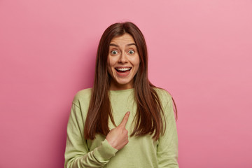 Obraz premium Portrait of surprised pleasant looking young girl indicates at herself, asks question, happy being chosen, cannot believe eyes, wears casual jumper, has long straight hair, isolated on pink background