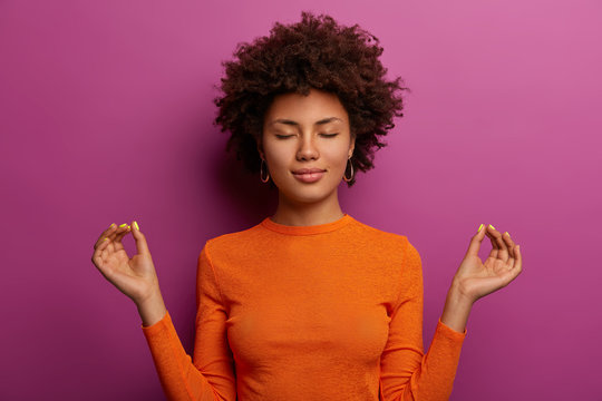 Peaceful Determined Curly Young Afro American Woman Makes Zen Gesture, Has Yoga Breathing Practice, Meditates Indoor, Closes Eyes And Wears Orange Jumper, Isolated Over Vibrant Purple Background