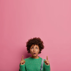 Confused shocked woman with Afro hairstyle, points upwards and says wow, promots product and...