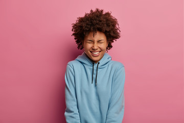Fototapeta na wymiar Photo of glad ethnic woman laughs and squints face, shows white teeth while smiles broadly, wears blue casual sweatshirt, poses against pastel rosy wall, expresses positive emotions and happiness