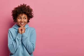 Fototapeta na wymiar Sincere positive dark skinned model holds hands under chin, being in good mood, wears blue sweatshirt, has happy smile on face, models against pastel rosy wall. People, emotions and lifestyle