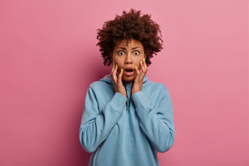 Fototapeta na wymiar Scared shocked Afro American woman stares with wide opened eyes, trembles from fear, drops jaw, hears bad news or rumors, wears blue hoodie, models over rosy pastel background. Human reaction