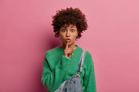 Shut up, please! Serious surprised curly woman demands silence, tells quiet and shushes with pressed index finger to lips, warns you about something, makes hush gesture, wears fashionable outfit