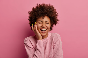Happy enthusiastic extremely positive woman touches cheek and laughs sincerely, stands delighted, closes eyes with pleasure, wears casual rosy pastel jumper, stands indoor, expresses good emotions.