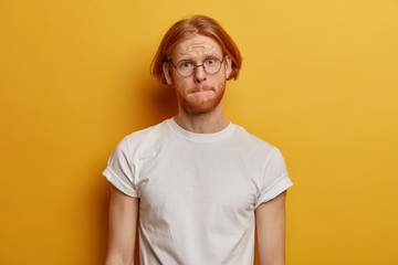 Handsome bearded man presses lips, looks with worried expressionn, tries to remember answer on question at exam, has trendy foxy hairstyle wears round glasses and casual outfit stands in yellow studio