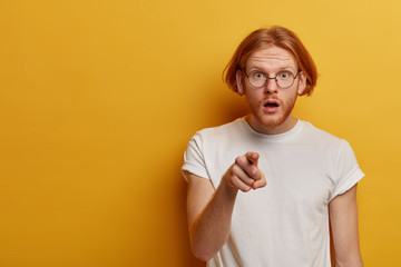 Shocked embarrassed bearded man with ginger hair and beard, points index finger at camera, has bated breath, impressed to notice someone in front, dressed casually, isolated on yellow studio wall