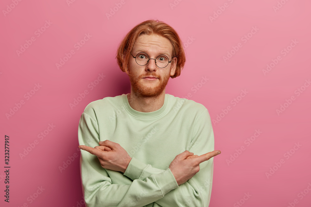 Wall mural Presentation, advertising, choice concept. Unsure bearded redhead man points reft and right corners, indicates sideways, chooses between two products, eager to choose something, dressed casually - Wall murals