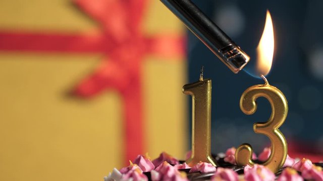 Birthday cake number 13 golden candles burning by lighter, blue background gift yellow box tied up with red ribbon. Close-up and slow motion