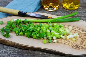 Close up of isolated sliced fresh spring onions (scallions) on wooden olive tree cutting board and japanese kitchen knife. Blue napkin, oil bottles and wood table background.