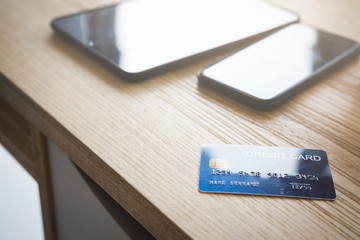 Blue credit card with smartphone and tablet on wooden table near window.