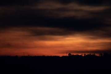 A silhouette of a line of trees and a power line in front of a dramatic red and orange sunrise in the far distance