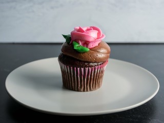 Chocolate cupcake with chocolate frosting topped with a pink frosting rose with green leaves on a plate with a gray and black background perfect for a birthday, love, Mother’s Day or Valentine’s Day.