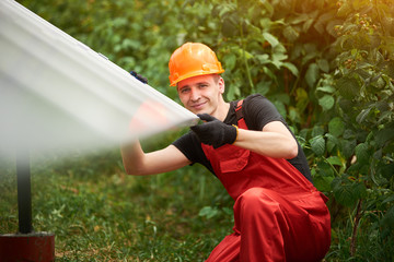 Happy man worker in orange uniform and safety helmet on blurry background of solar panels and greenery. Home construction of solar station. Alternative energy sustainable concept