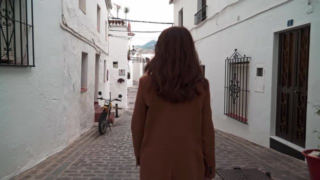 Zoom in gimbal shot of back of woman dressed in brown coat walking in narrow white street in old city Mijas, a municipality in the Province of Malaga, in winter time