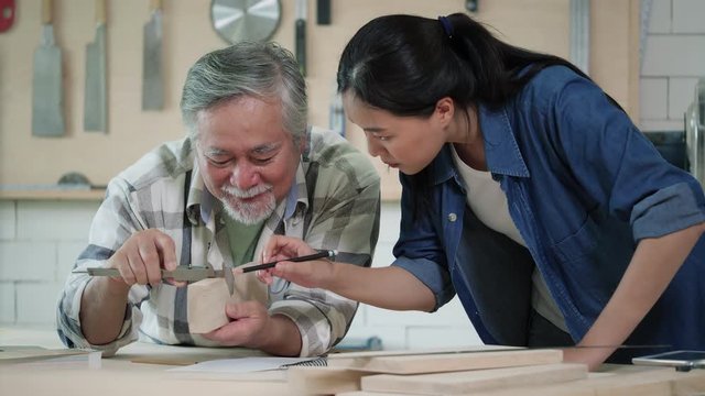Asian senior male and teenage girl carpenter working design modern furniture in home workshop. Carpenter is high experience and presentation skill in lumber.